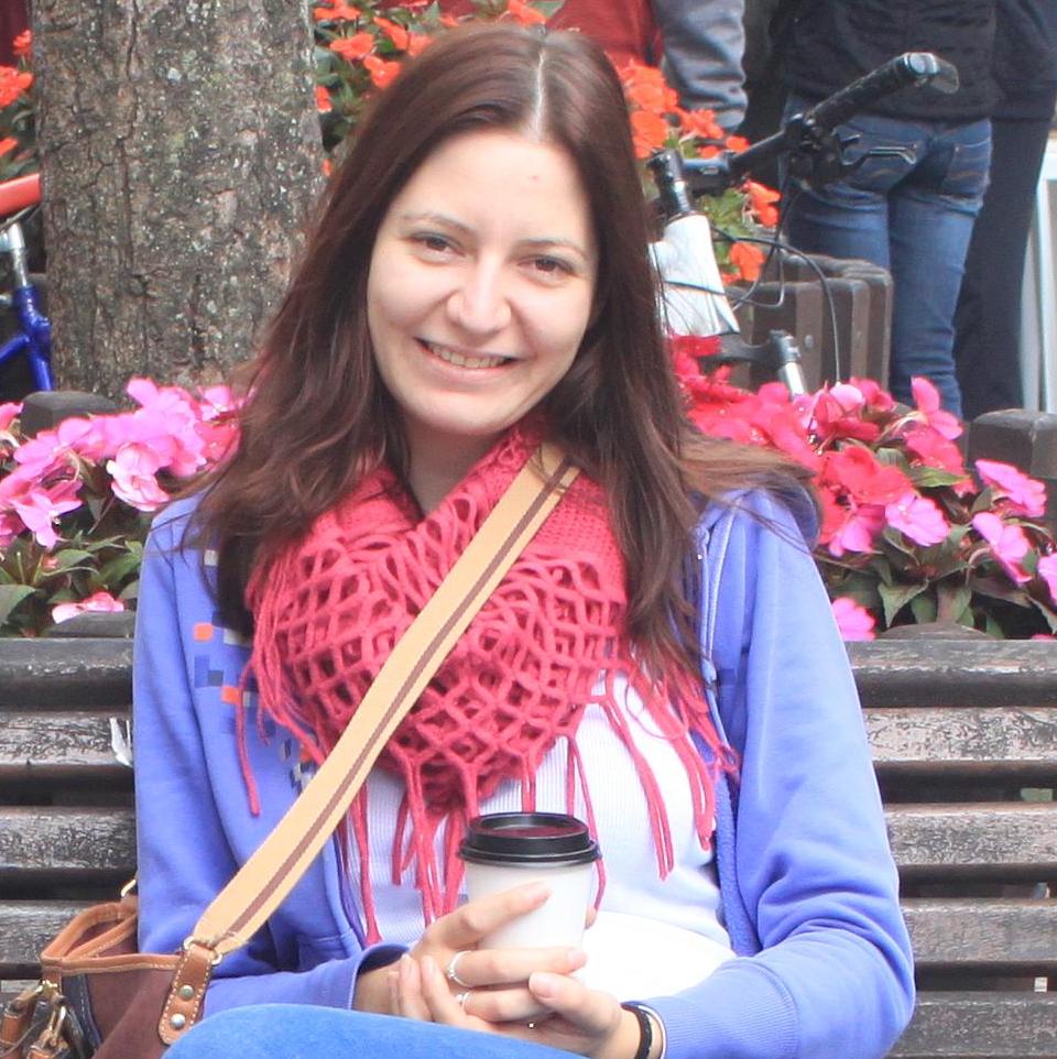 Aqui é uma foto da pesquisadora Ana Paula, caucasiana, cabelo claro, sentada em uma praça com um copo de café na mão. Ela está sorrindo