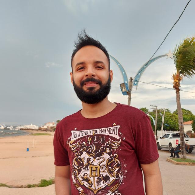 Aqui é uma foto do pesquisador Davi sorrindo. É homem, pardo, de barba e cabelos escuros, está com uma camiseta do harry potter e está na frente de um rio.